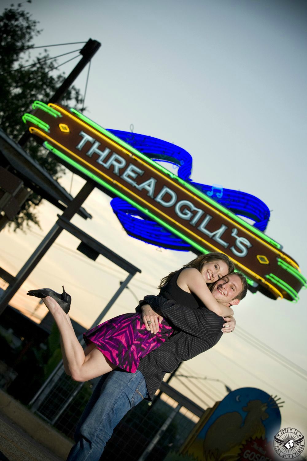 Dirty blond haired girl wearing a black and pink dress and black heels jumps into the arms of her fiancé, a guy with dishwater blond hair wearing a long sleeve black button up shirt and blue jeans in front of the Threadgill's sign with a blue and orange sky in this colorful engagement snapshot in downtown Austin.